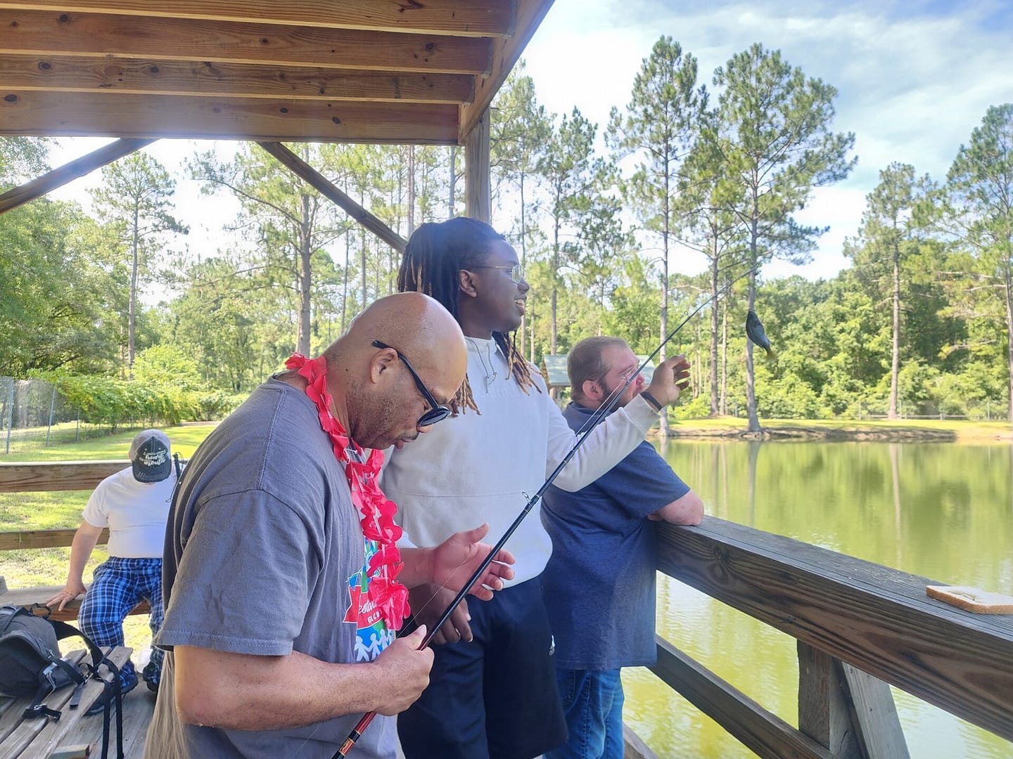 Four people are fishing from a wooden structure by a serene, tree-lined pond. The foreground shows three individuals closely engaged in fishing; one adjusts his rod, another inspects the line, and a third looks towards the water. This bright and sunny scene perfectly captures moments from summer programs.