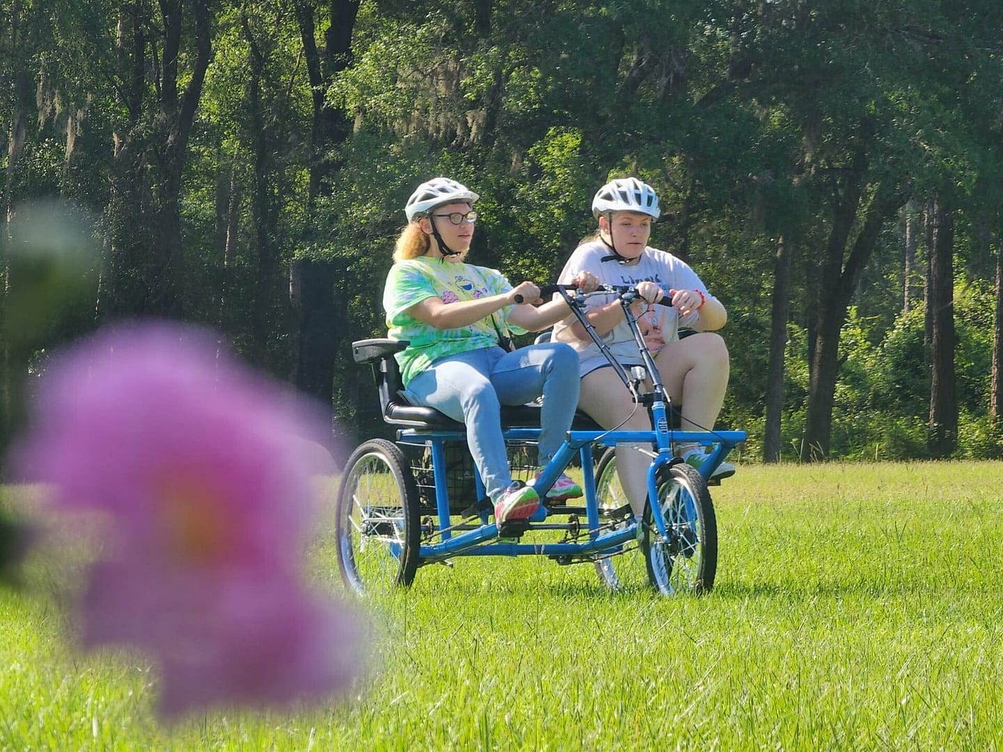 Two people are riding a blue tandem bicycle on a grassy field surrounded by trees, like participants in one of those outdoor wellness programs. Both are wearing white helmets and casual clothing. The foreground shows a blurred pink flower, adding a touch of color to the scene.