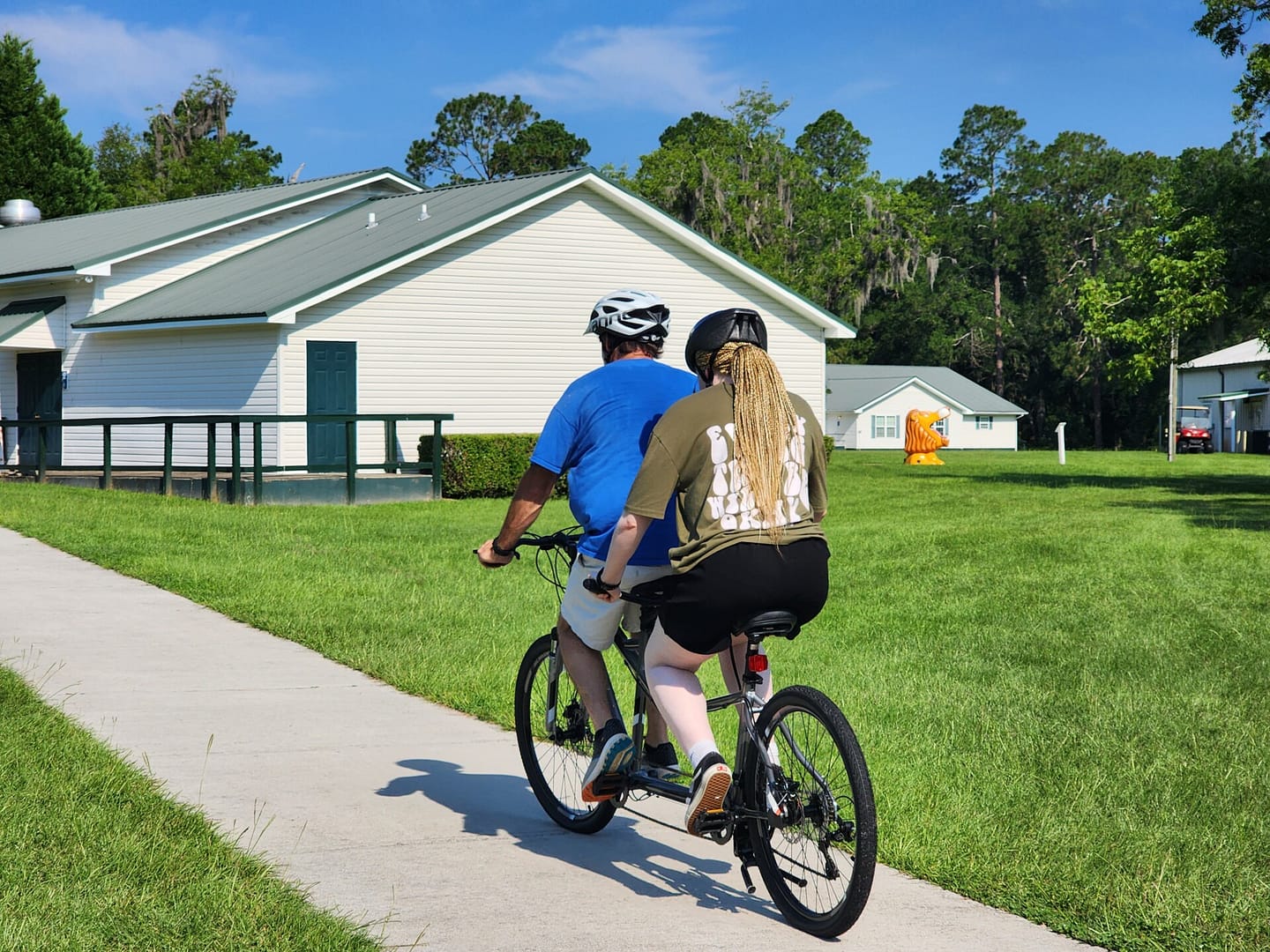 Two people wearing helmets ride a tandem bicycle on a paved path through a grassy area, reminiscent of leisurely summer programs. White buildings with green roofs and a small orange dinosaur sculpture grace the background, while trees and a clear blue sky complete the scene.