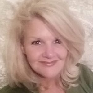 A woman with shoulder-length blond hair is smiling softly at the camera. She has light-colored eyes and is wearing a green top. The background showcases a light, patterned surface that complements her serene expression perfectly.