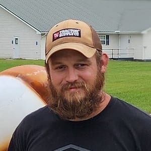 A bearded man wearing a tan "Tractor Supply Co." cap and a black shirt stands outside in front of a grassy area with a white building about 20 feet in the background.