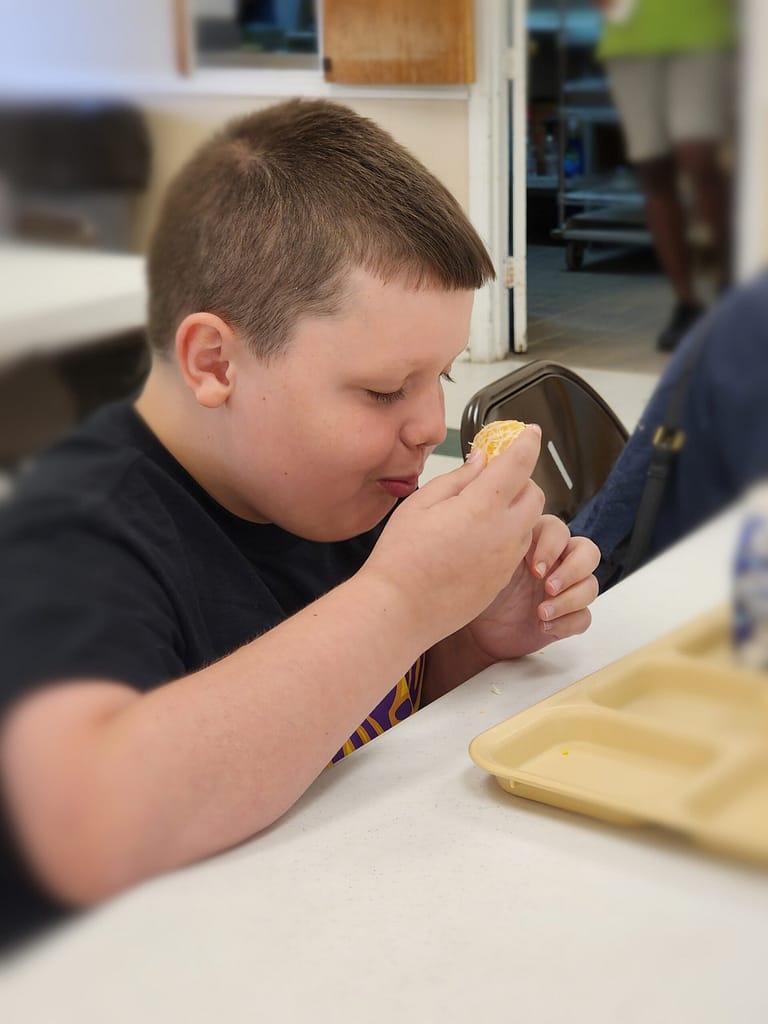 A young boy with short brown hair sits at a table in a cafeteria, wearing a black shirt. He is holding an orange that he just peeled close to his mouth, ready to take a bite. In front of him is an empty beige cafeteria tray. The background shows blurred people and furniture.