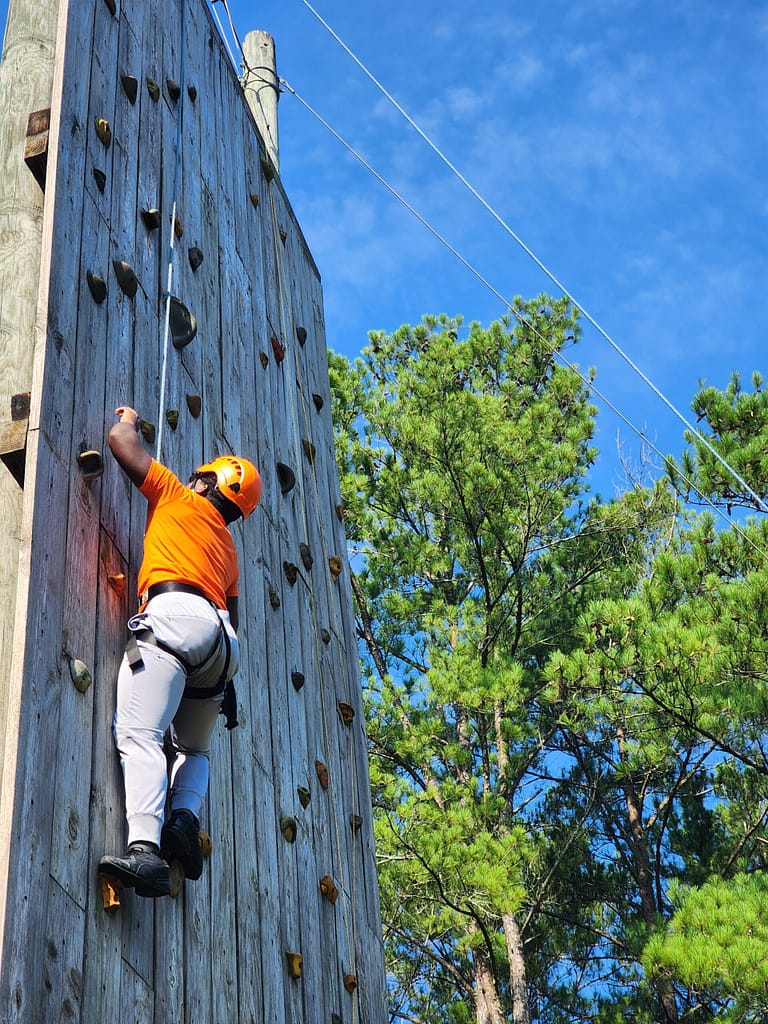 A person wearing an orange helmet and orange shirt climbs a vertical wooden rock climbing wall. The sky is clear and blue, with green trees in the background. The person skillfully uses hand and footholds for support.